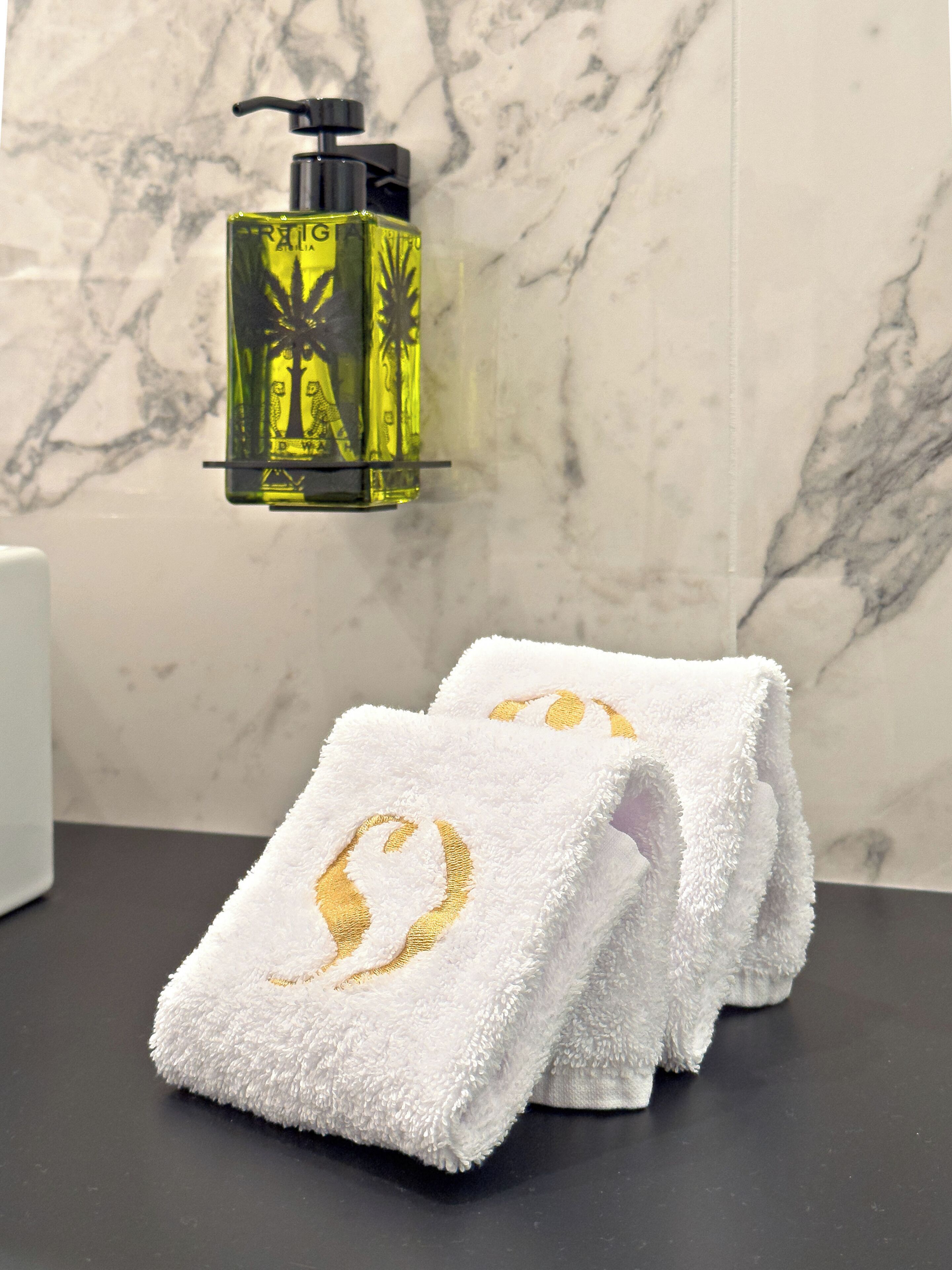 A bathroom with a marble wall, a yellow bottle of liquid soap, and two white towels with golden embroidery.