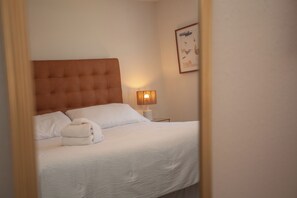 Una cama bien tendida con sábanas blancas, cabecera de madera y lámpara de noche.