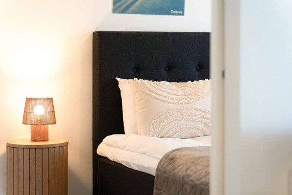 Un lit bien fait, avec une literie blanche et des coussins décoratifs, une table de chevet ronde en bois avec une lampe, et un panneau décoratif fixé au mur au-dessus du lit.