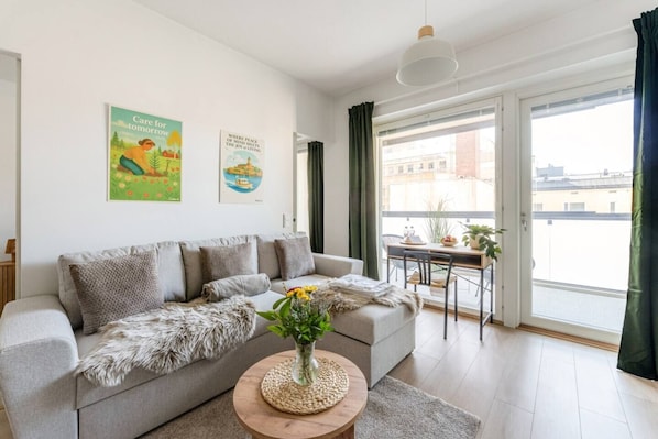 Una sala de estar moderna con un sofá gris, una mesa redonda de centro con un florero, un comedor con mesa y sillas, y un gran ventanal con cortinas.
