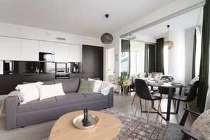 Un saló modern amb un sofà gris, una zona de menjador amb una taula rodona i una cuina amb armaris blancs.