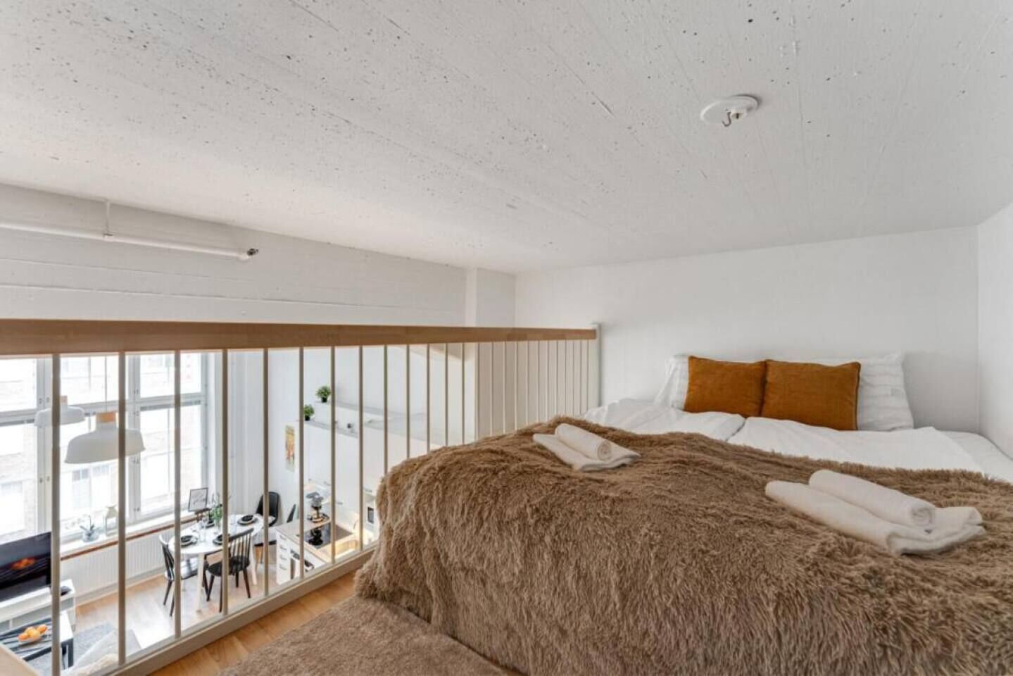 A bedroom with a large bed, a view of the city through floor-to-ceiling windows, and a dining area visible through the windows.