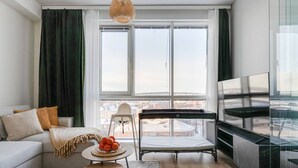 A modern living room with a sofa, a small table with fruit, a baby chair, a balcony with a mattress, and a TV. - Arena Comfort Queen-Free Gym-Views (Tampere)