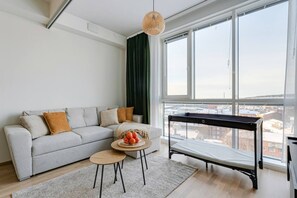 A modern living room with a sofa, a small round table with fruit, a standing bed, and large windows offering a city view. - Arena Comfort Queen-Free Gym-Views (Tampere)