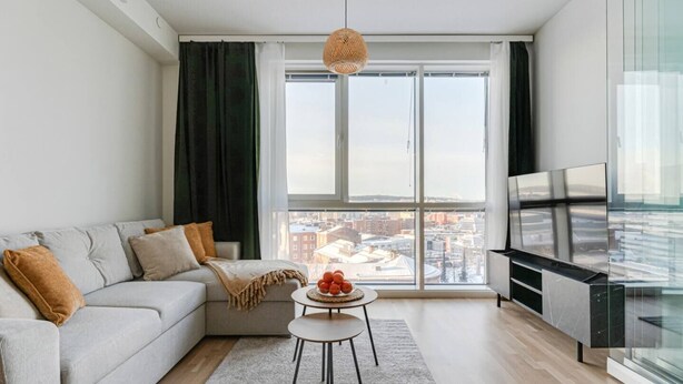 A modern living room with a sofa, a small round table with fruit, a TV, and a large window with a city view. - Arena Comfort Queen-Free Gym-Views (Tampere)
