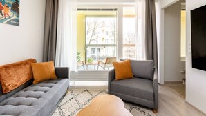 A modern living room with a grey sofa, a grey armchair, and a wooden coffee table. There is a large window with curtains, a view of buildings outside, and a flat-screen TV mounted on the wall.