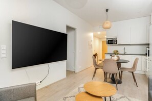 Moderni olohuone, jossa on suuri litteänäyttöinen televisio, ruokailutila pyöreällä pöydällä ja tuoleilla sekä keittiö, jossa on valkoiset kaapit ja mikroaaltouuni.