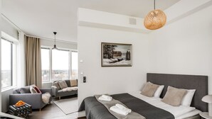 A modern hotel room with a large bed, a sofa, a dining table, and a city view.