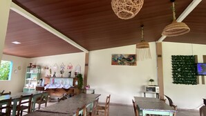 A dining area with wooden tables and chairs, a hanging woven light fixture, a wall-mounted painting, and a television.