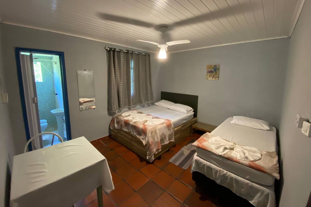 A room with two beds, a table, a ceiling fan, and a door leading to a bathroom.