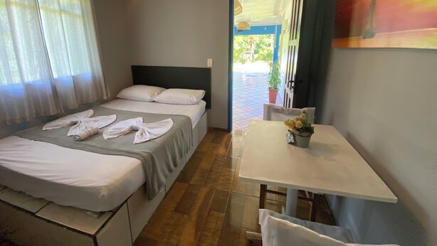 A room with a bed, a table, and a chair. There is a window with curtains and a door leading outside.