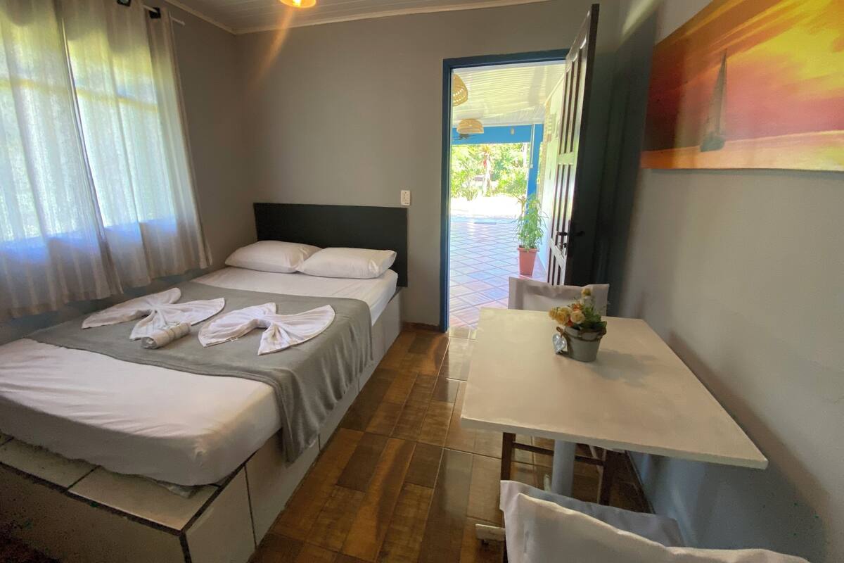 A room with a bed, a table, and a chair. There is a window with curtains and a door leading outside.