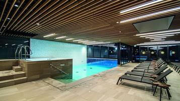 En moderne swimmingpool indendørs med træloft, glasvæg og liggestole.