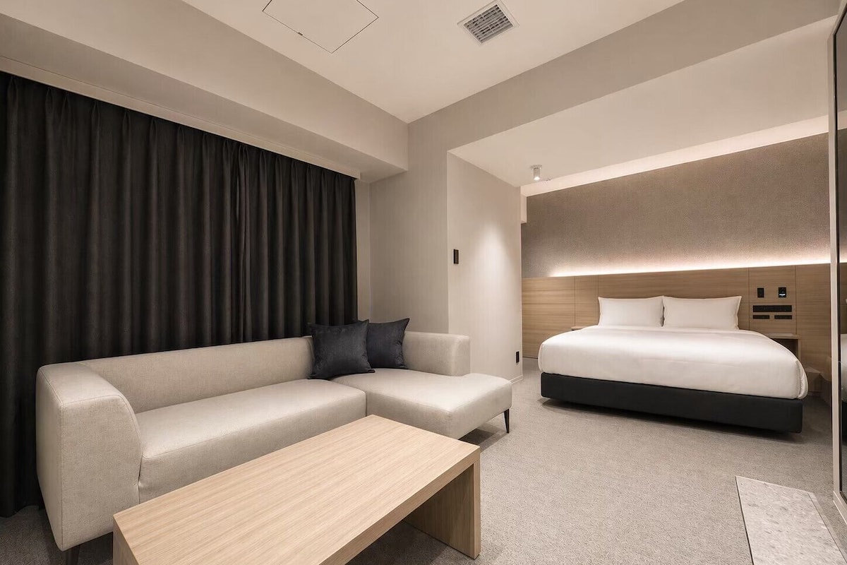 A modern hotel room with a sectional sofa, a single bed, a wooden coffee table, and a large window with curtains.