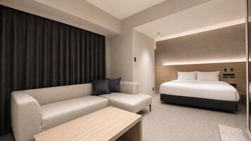 A modern hotel room with a sectional sofa, a single bed, a wooden coffee table, and a large window with curtains.