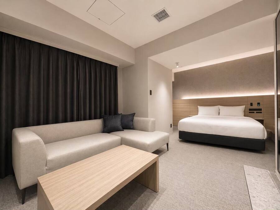 A modern hotel room with a sectional sofa, a single bed, a wooden coffee table, and a large window with curtains.