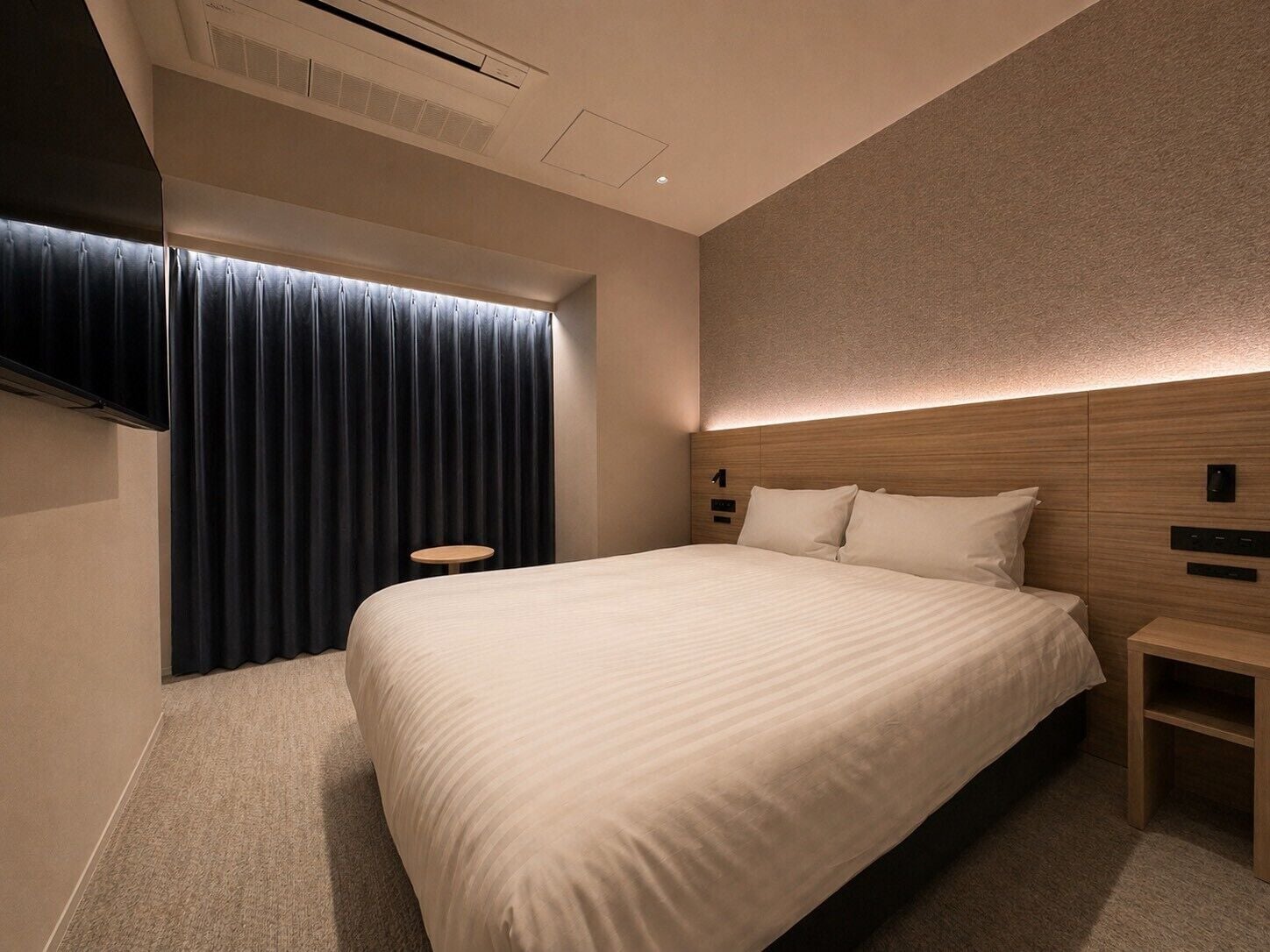 A hotel room with a large bed, a bedside table, a wall-mounted TV, and a window with curtains.