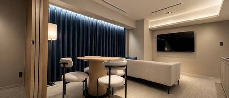 A modern hotel room with a round table, four chairs, a sofa, a TV, and a large window with curtains.