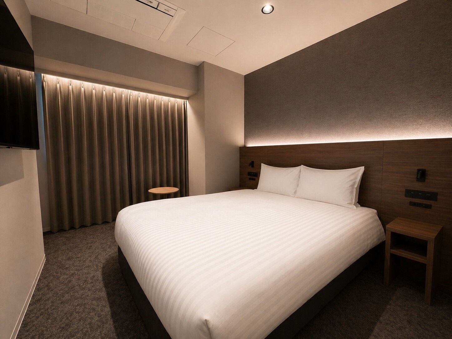A hotel room with a large bed, a TV mounted on the wall, a bedside table, and a curtain.