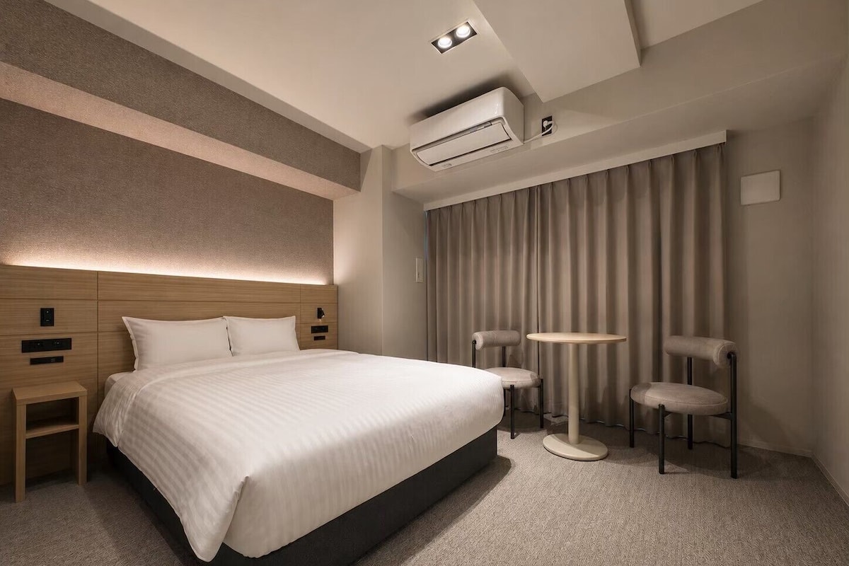 A modern hotel room with a large bed, a bedside table, a round table, two chairs, and an air conditioning unit.