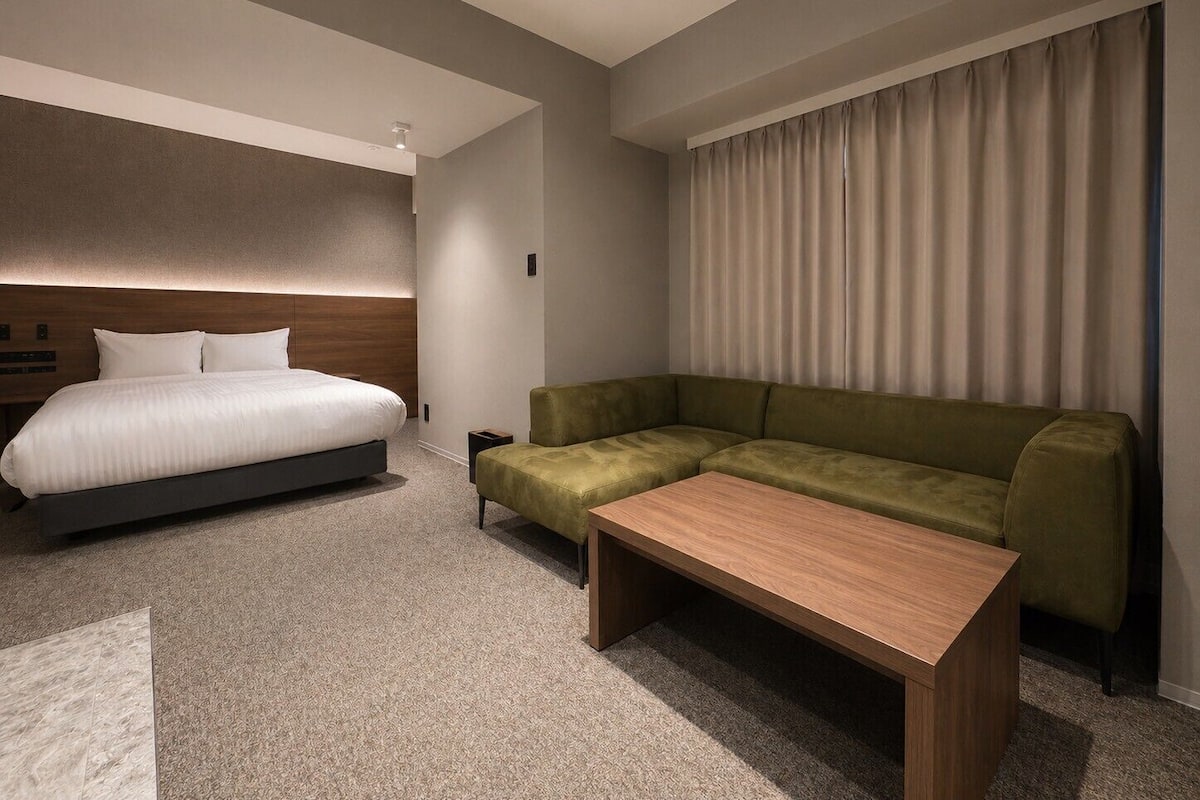 A modern hotel room with a bed, a green sofa, a wooden coffee table, and a large window with curtains.