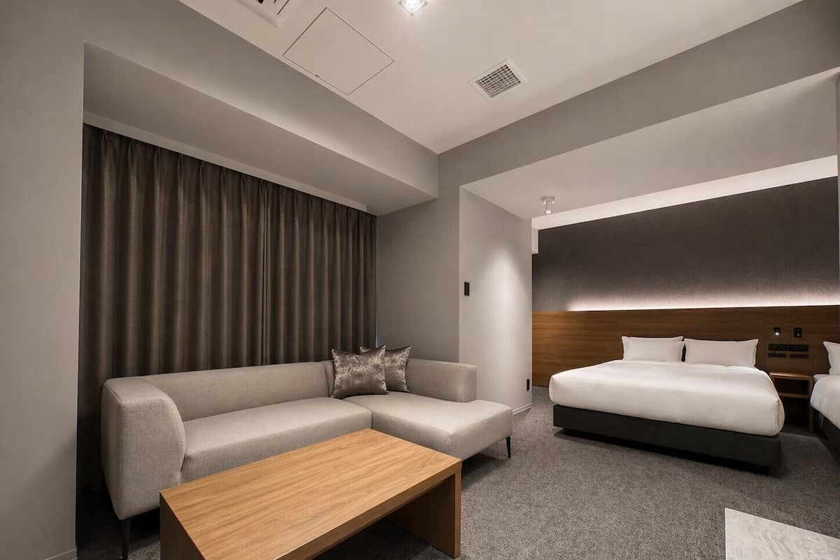 A modern hotel room with a sofa, a wooden coffee table, a bed, and a headboard with built-in lighting.