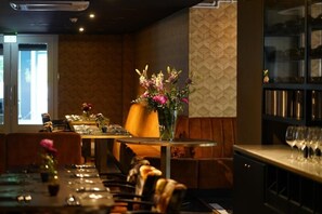 A restaurant with a long table set for dining, a bar area with glasses, and a decorative vase with flowers.