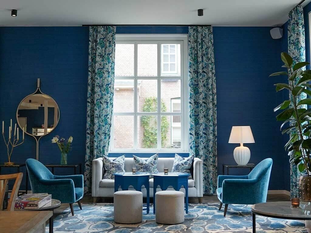 A room with a large window, blue walls, a white sofa, two blue armchairs, a small table, and a lamp.