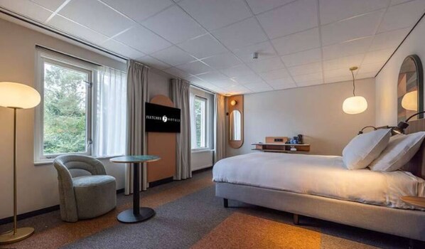 Une chambre d’hôtel avec un lit, une chaise, une petite table, une télévision et de grandes fenêtres.