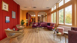 Un hall d’hôtel avec un plancher en bois, des murs rouges et un canapé violet. On y trouve des chaises en osier et une petite table sur laquelle est posé un vase.