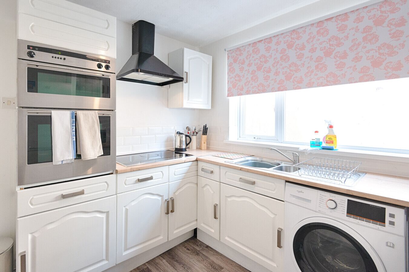A compact kitchen with a built-in oven, microwave, and washing machine, white cabinetry, and a window with floral blinds.