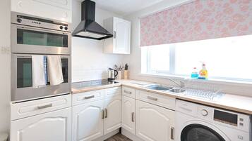 A compact kitchen with a built-in oven, microwave, and washing machine, white cabinetry, and a window with floral blinds.