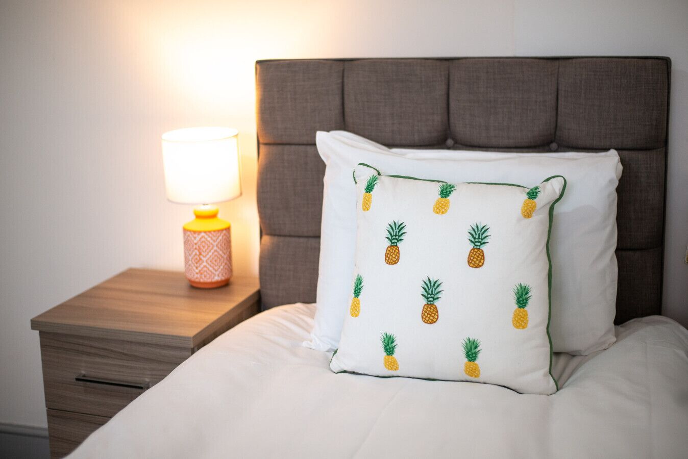 Ein ordentlich bezogenes Bett mit einem weißen Kissen mit Ananas-Motiven, ein Nachttisch aus Holz mit einer Lampe und ein dunkles, gepolstertes Kopfteil.