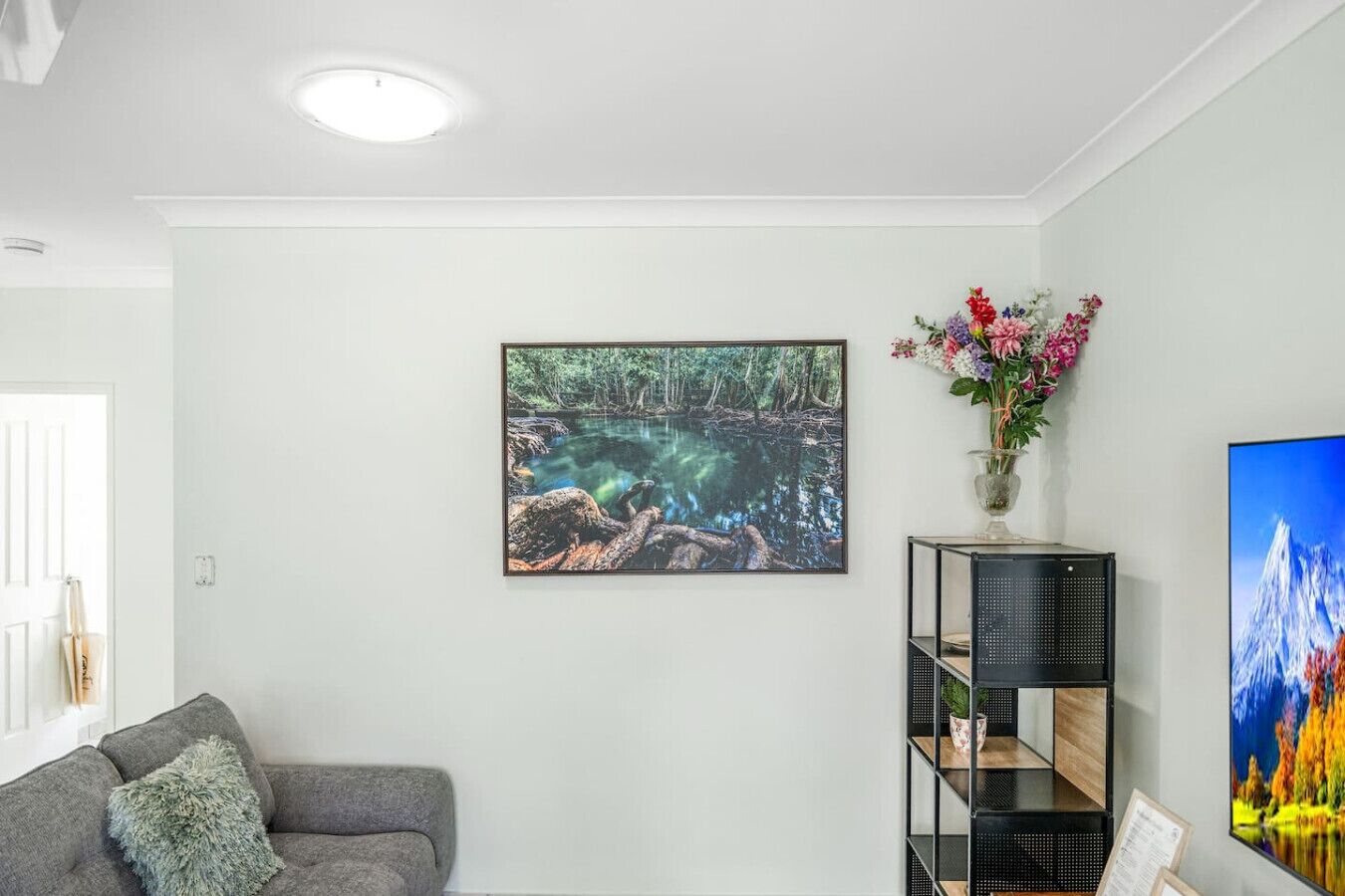 Una sala de estar con un sofá gris, una escena natural enmarcada, un mueble negro para estantería y un jarrón con flores.