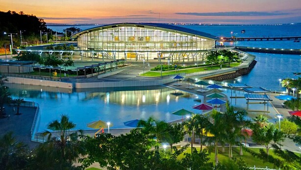 A modern building with a glass facade, surrounded by a waterfront promenade with umbrellas and palm trees.