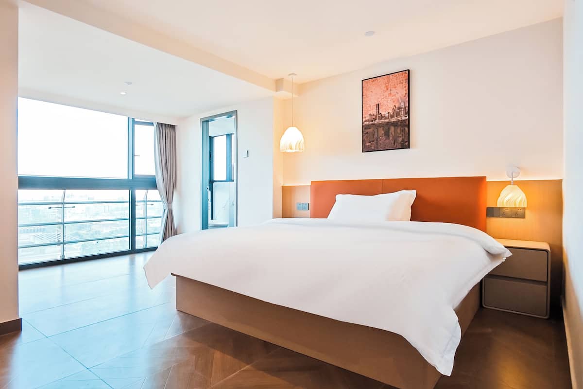 A hotel room with a large bed, a nightstand with a lamp, a cityscape painting on the wall, and a balcony with a view.
