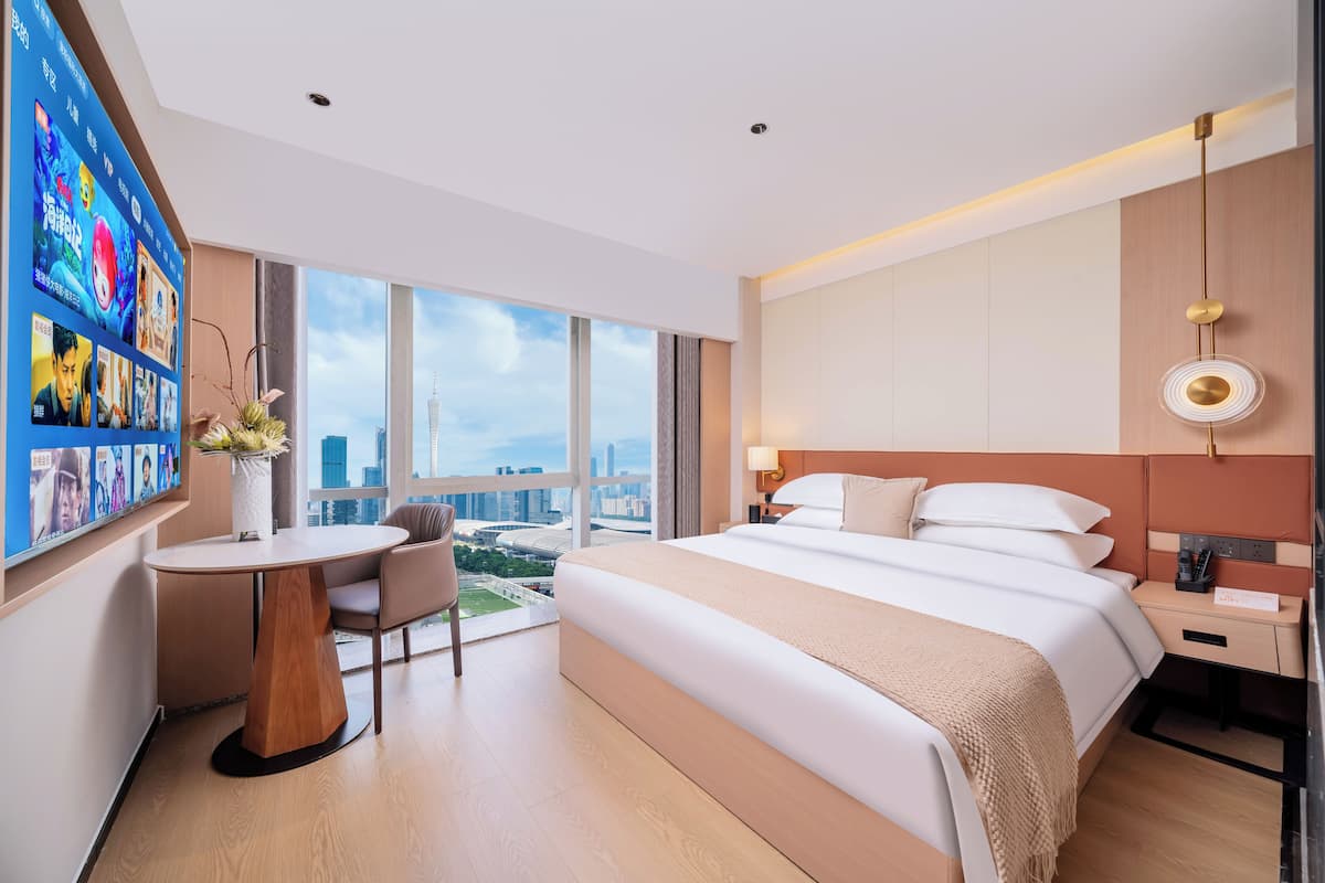A hotel room with a large bed, a desk, a chair, a TV on the wall, and a city view.