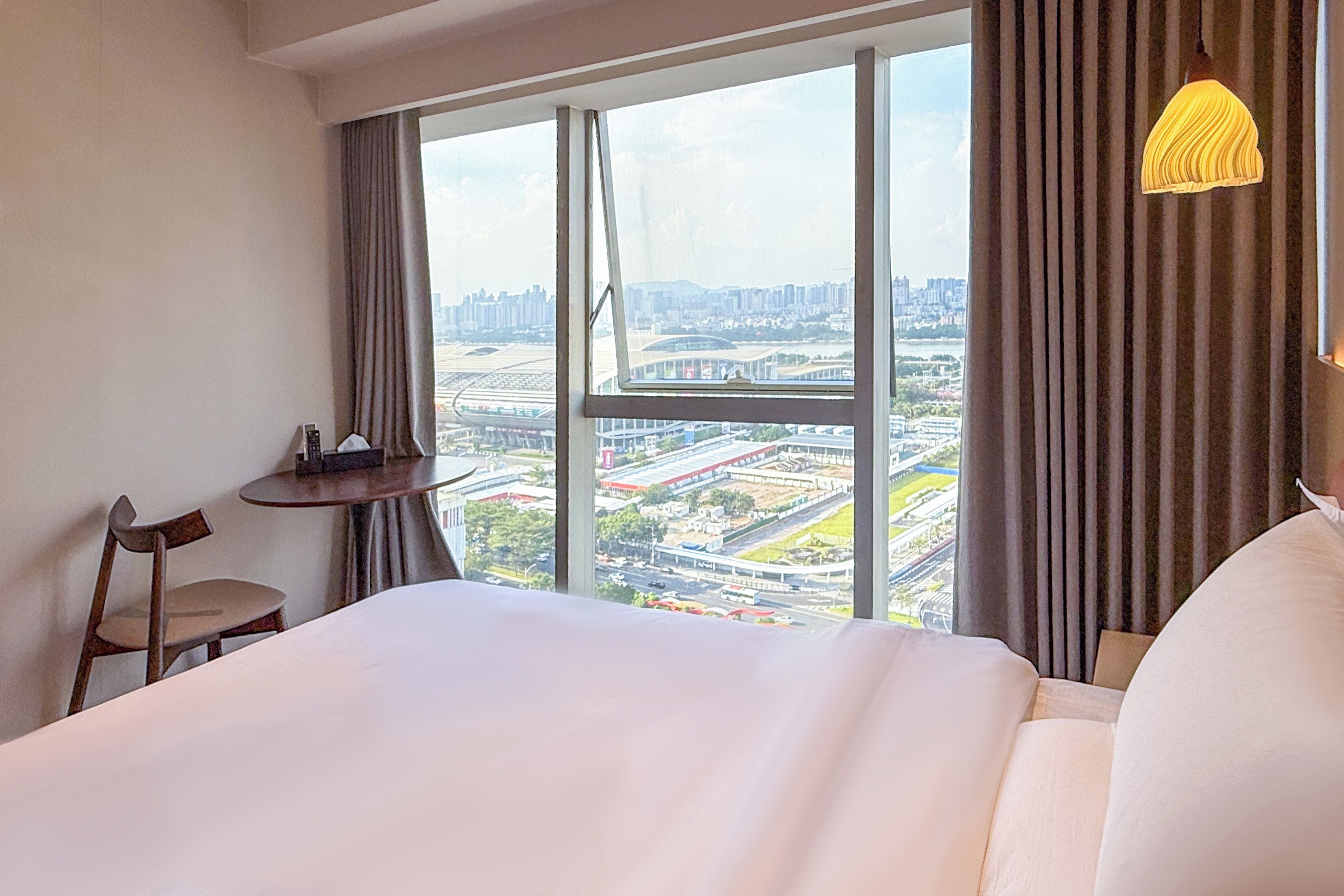A hotel room with a large bed, a small table, a chair, and a view of a cityscape through large windows.