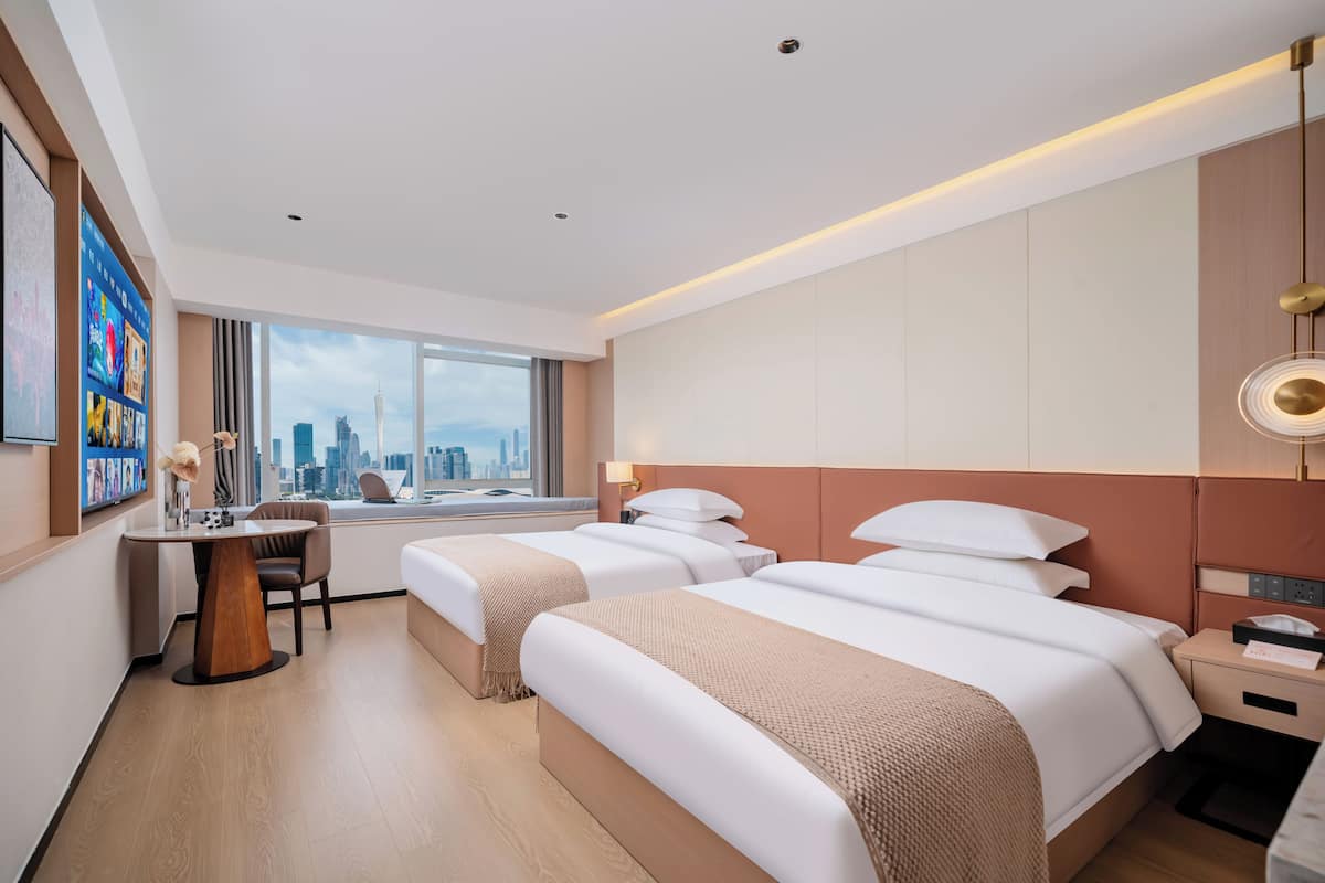 A hotel room with two beds, a desk, a chair, a window with a city view, and a wall-mounted TV.