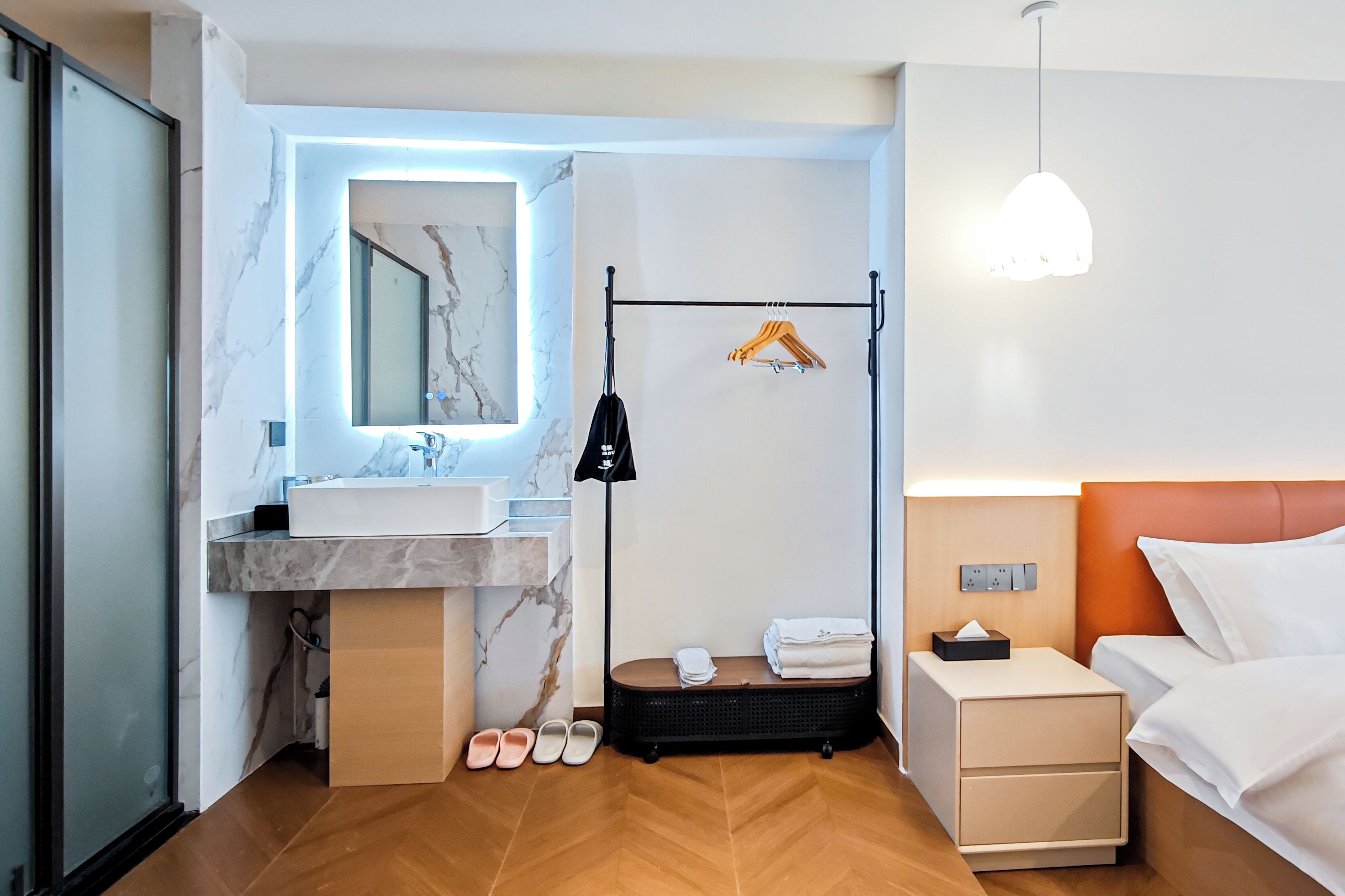 A modern hotel room with a marble bathroom vanity, a hanging clothes rack, a suitcase, and a neatly made bed.