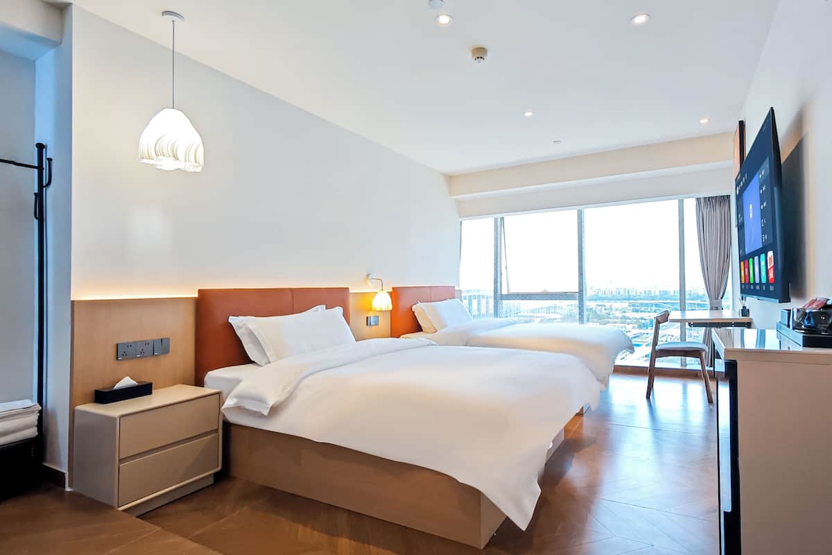 A hotel room with two beds, a desk, a TV, and a large window with a city view.