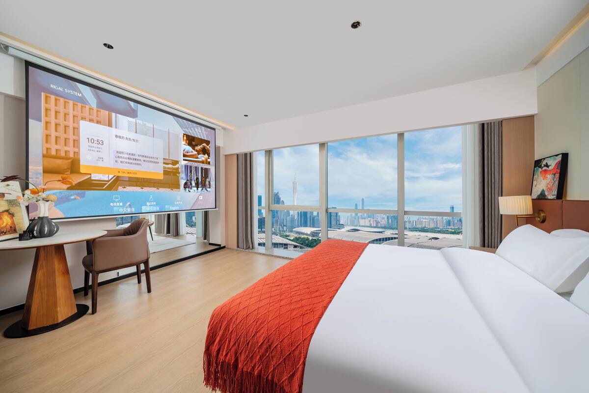 A hotel room with a bed, a desk, a chair, a TV, and a large window with a city view.