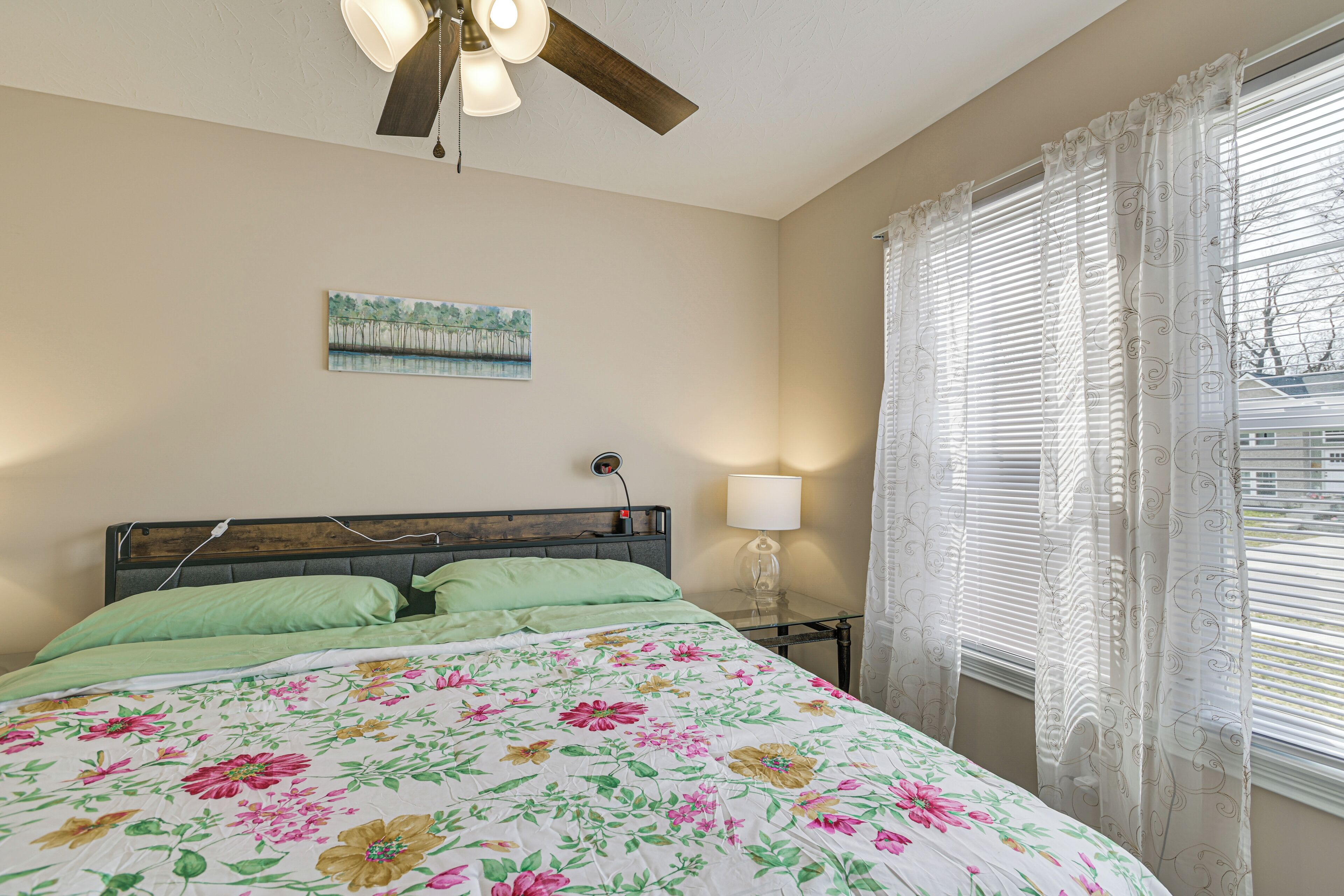 Une chambre à coucher avec un lit, un ventilateur de plafond, une table de chevet avec une lampe, un tableau au mur et une fenêtre avec des stores.