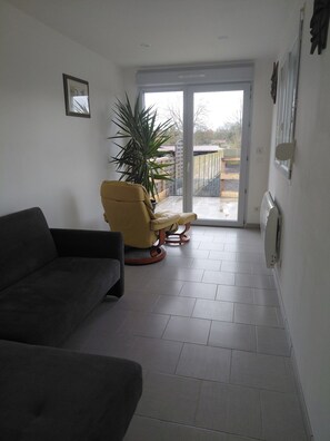 A living room with a sofa, a chair, a potted plant, and a sliding glass door leading to an outdoor area. - Town house in a simple commune. (WATTEN)