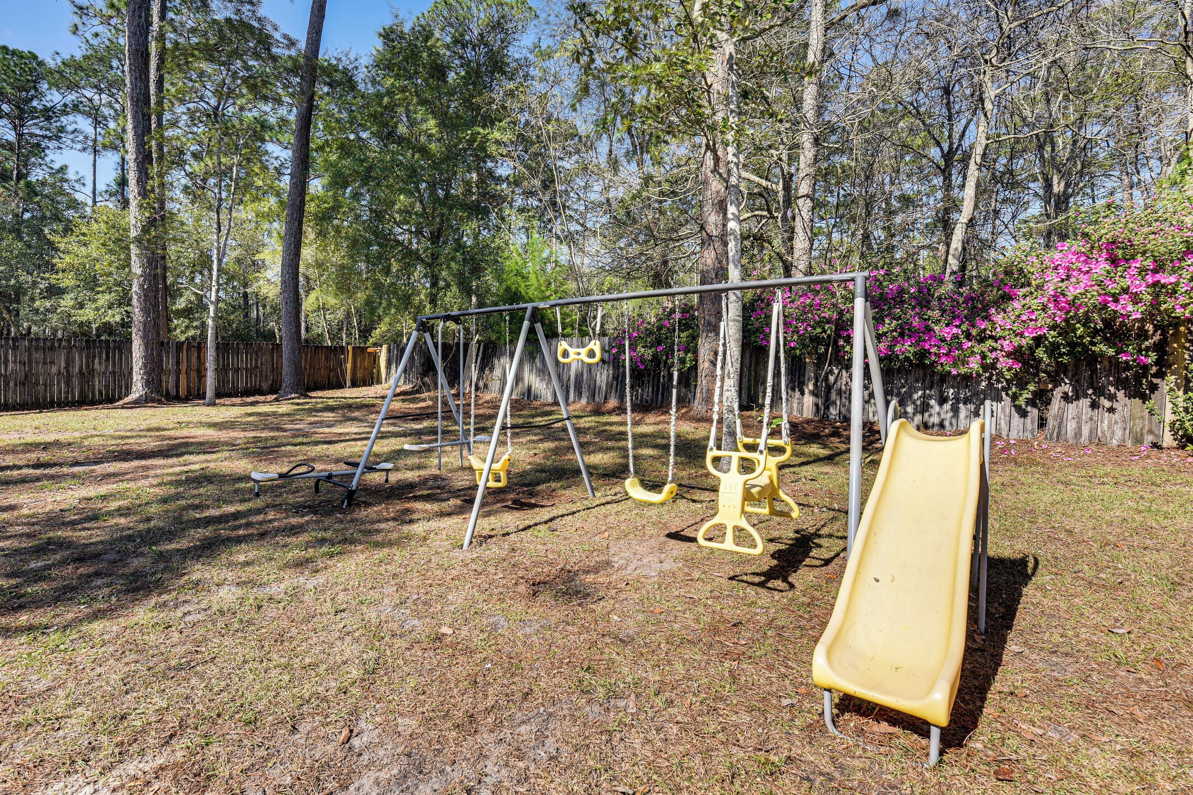 A playground with swings and a slide, surrounded by trees and a wooden fence.