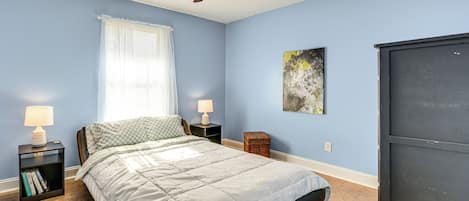 Un dormitorio con cama, ventilador de techo, mesita de noche con lámpara, ventana con persianas y una obra de arte colgada en la pared.