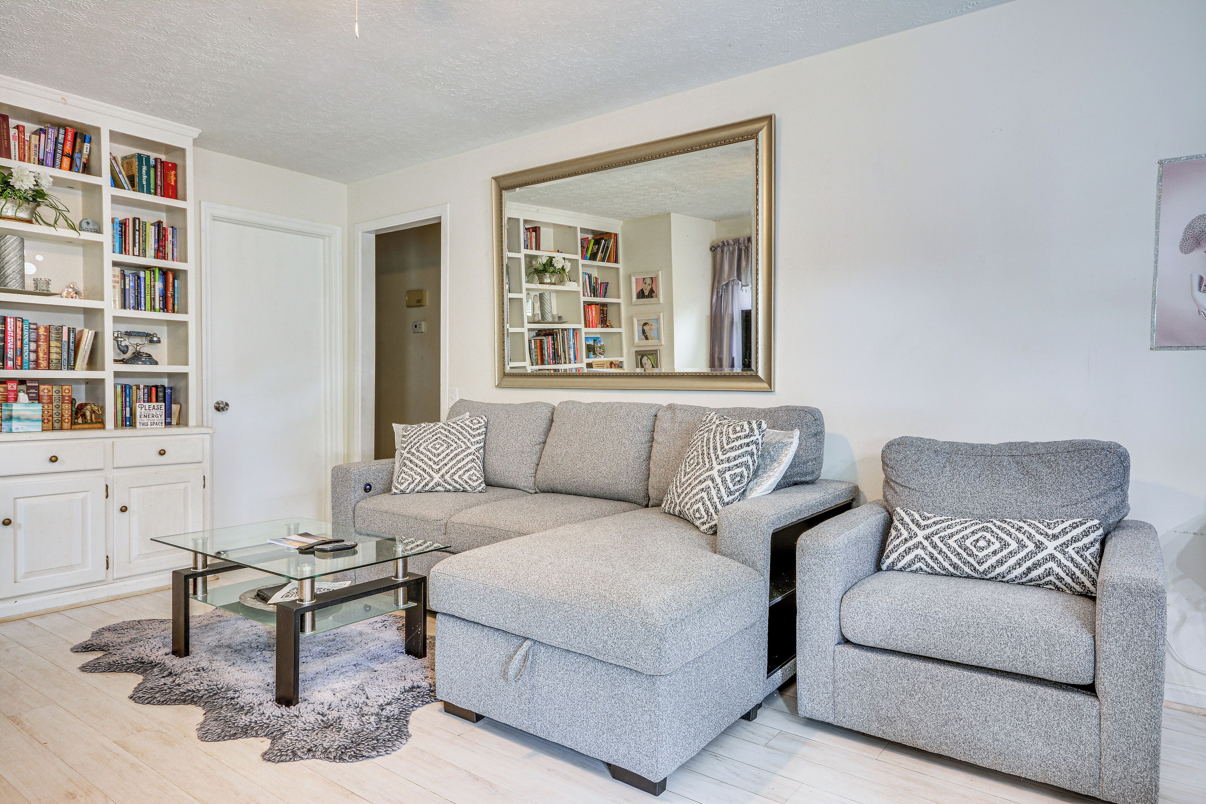 A living room with a sectional sofa, a glass coffee table, a bookshelf, and a large mirror.