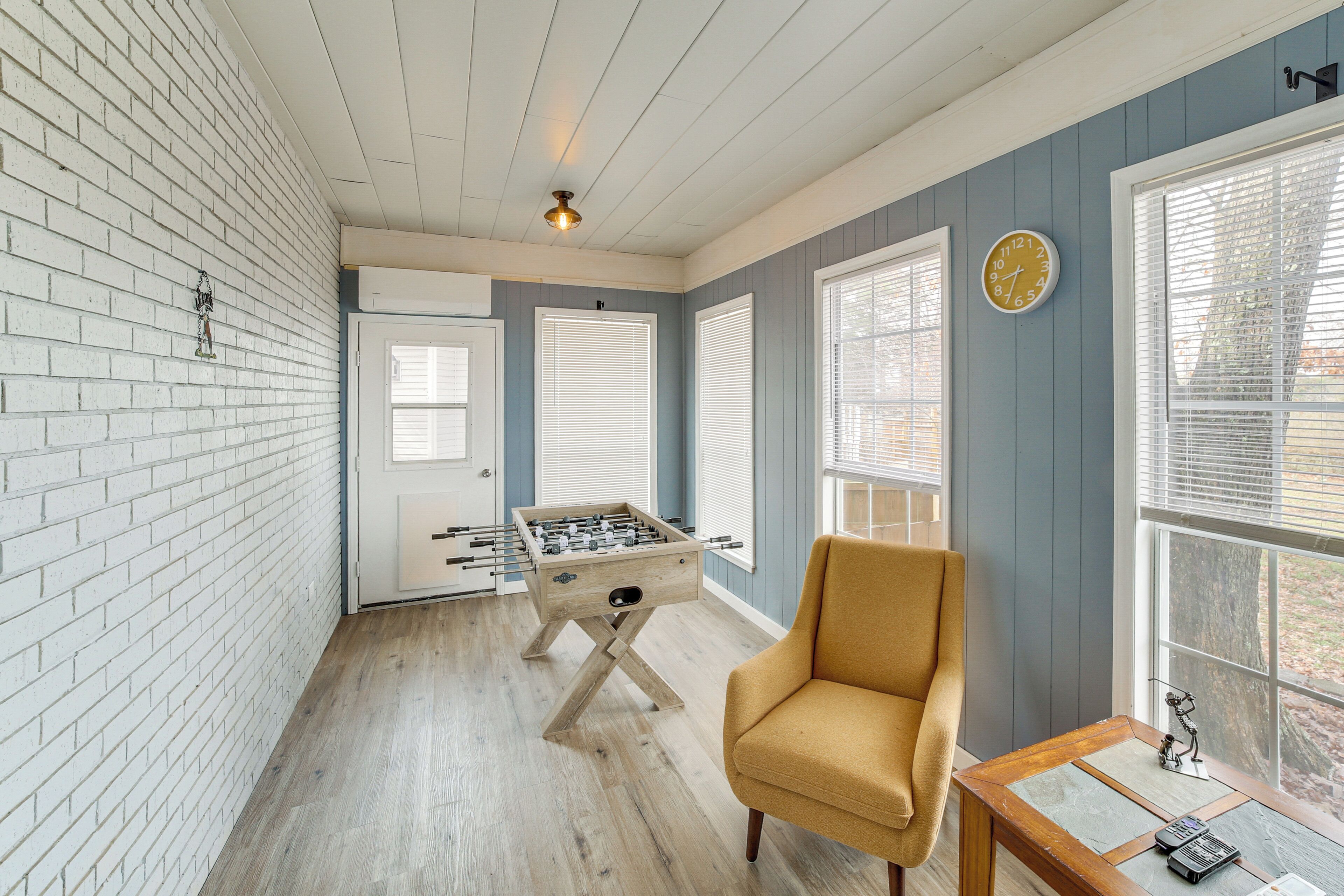 Une pièce avec un mur en briques, un plancher en bois, une table de baby-foot, un fauteuil jaune et une fenêtre avec des stores.