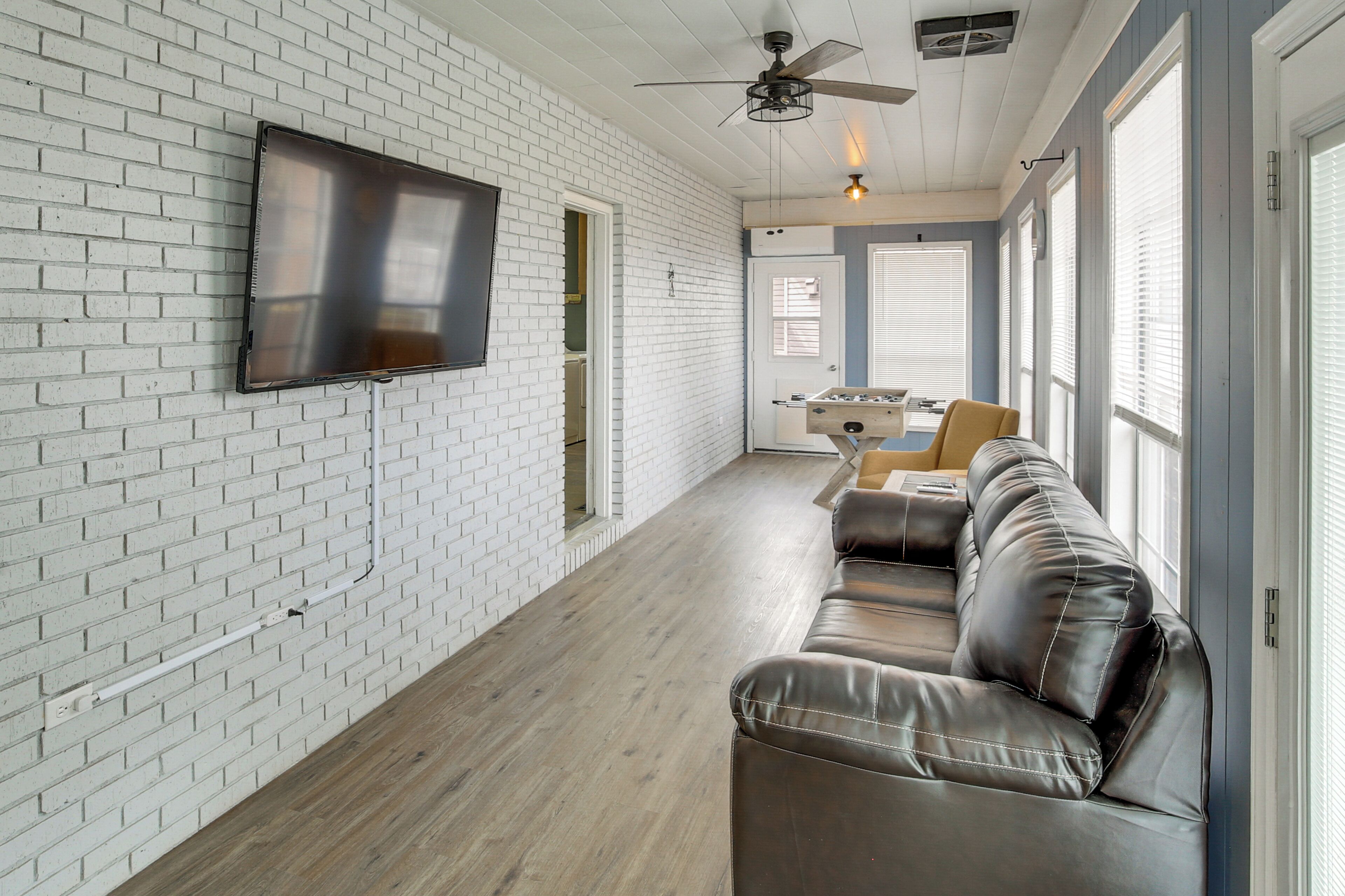 A modern living room with a white brick wall, a flat-screen TV, a ceiling fan, and a leather sofa.