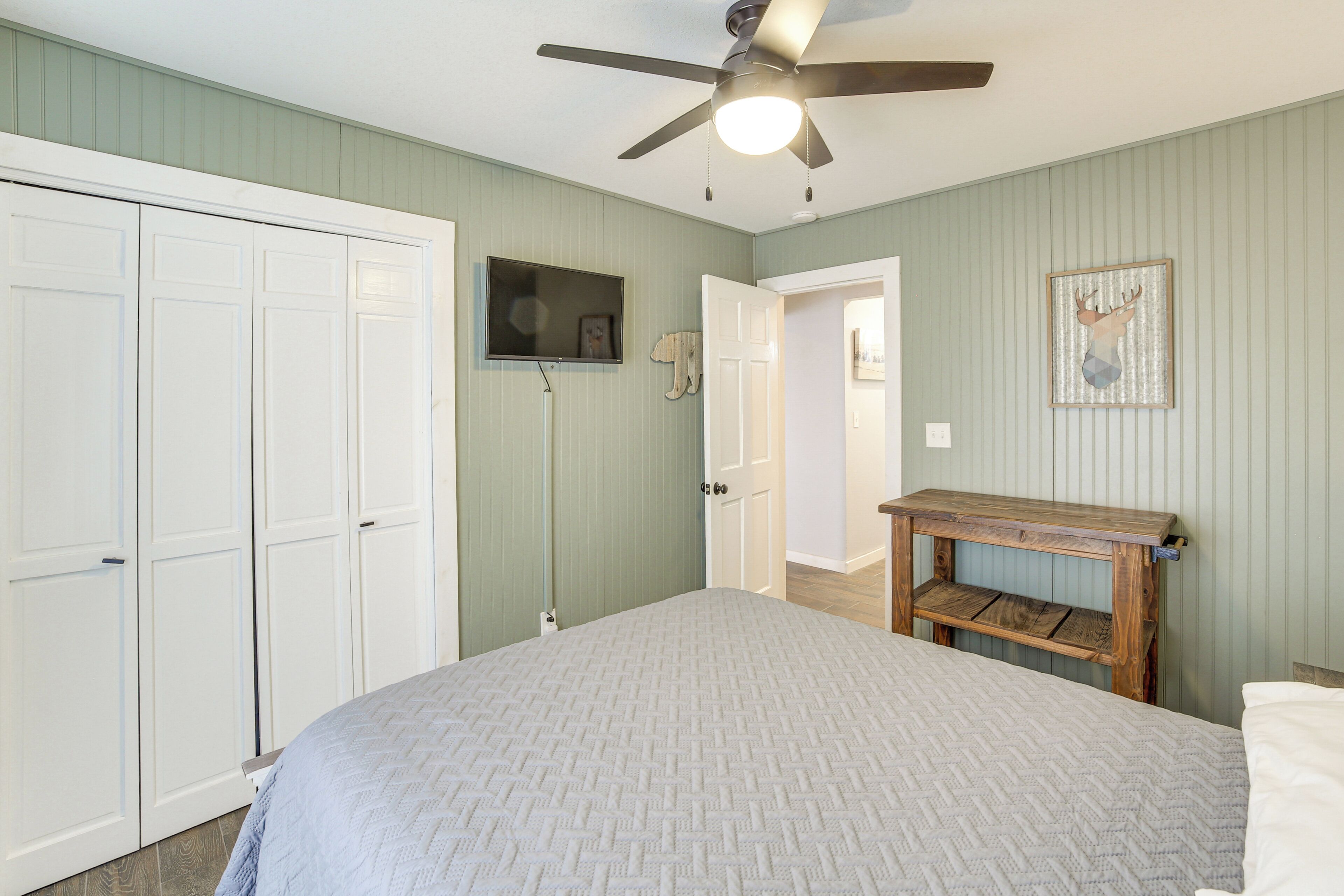 A bedroom with a bed, a ceiling fan, a TV, and a door leading to another room.